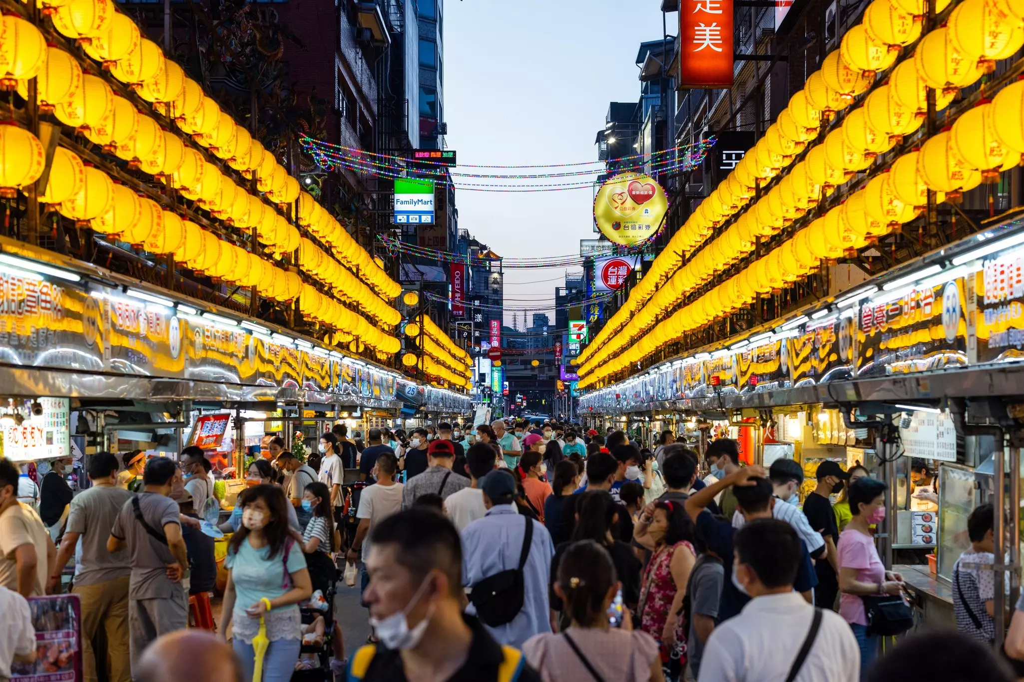 Hundreds of yellow lanterns illuminate a narrow street at night, crowded with food vendors and patrons.