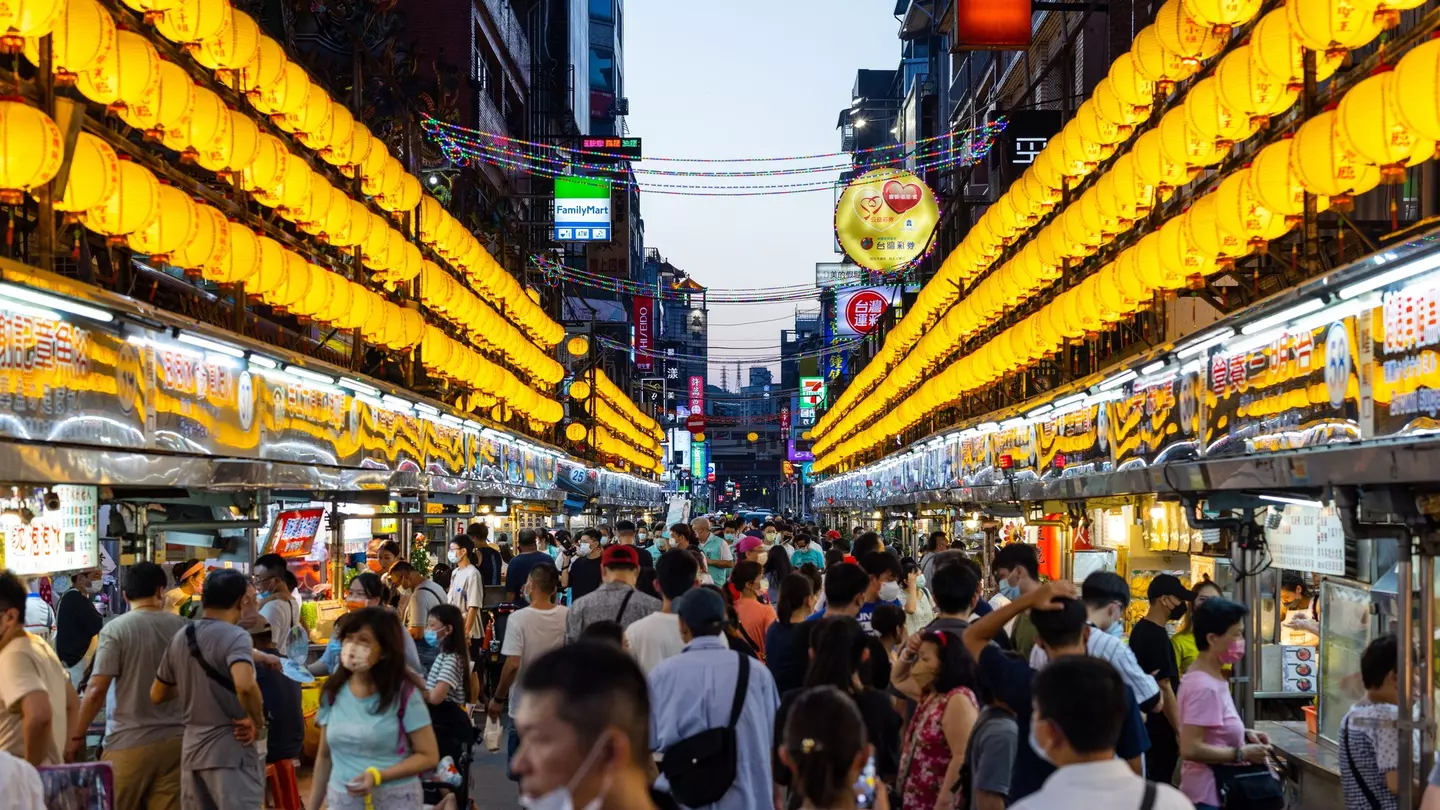 Hundreds of yellow lanterns illuminate a narrow street at night, crowded with food vendors and patrons.