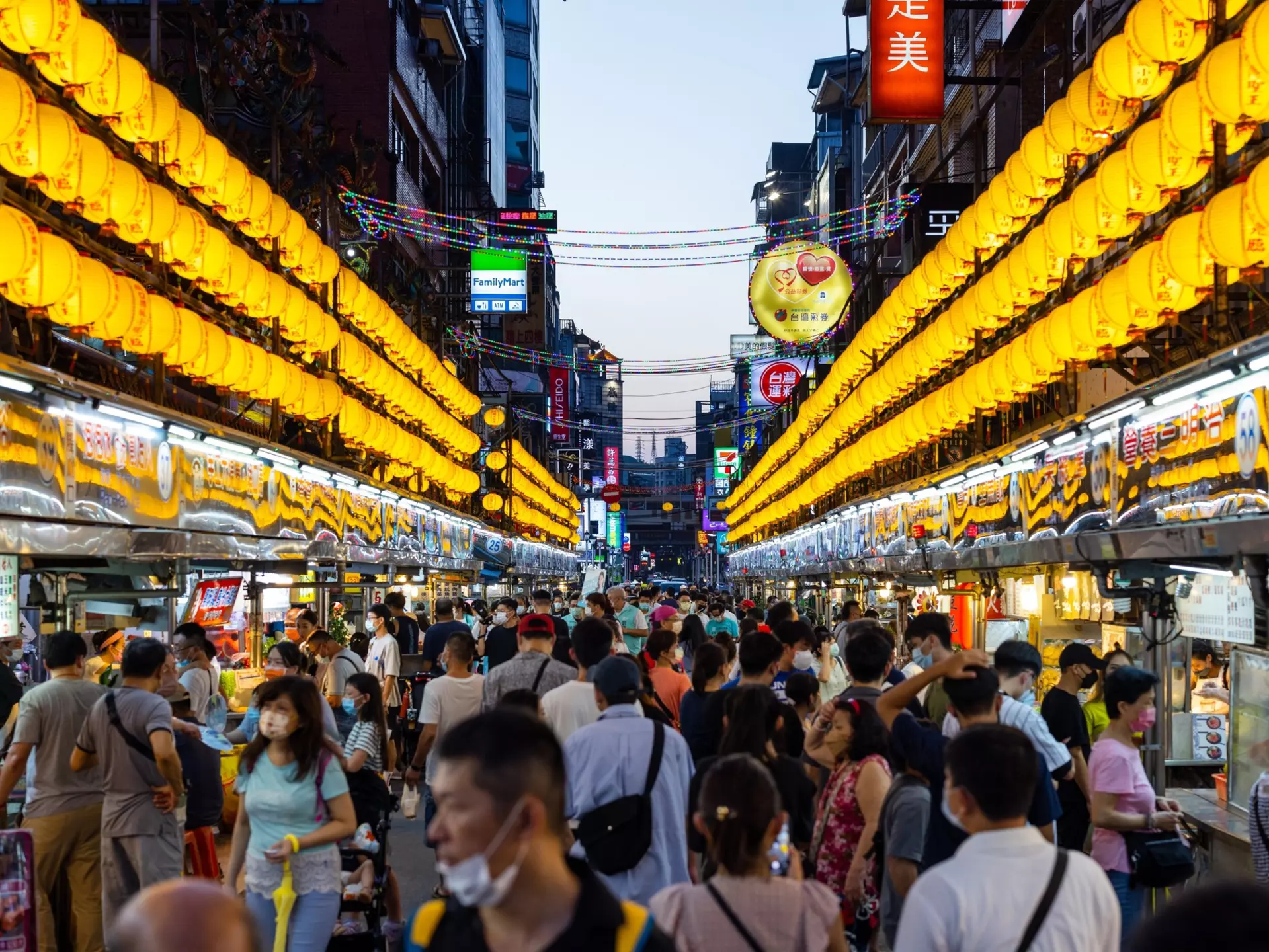 Hundreds of yellow lanterns illuminate a narrow street at night, crowded with food vendors and patrons.