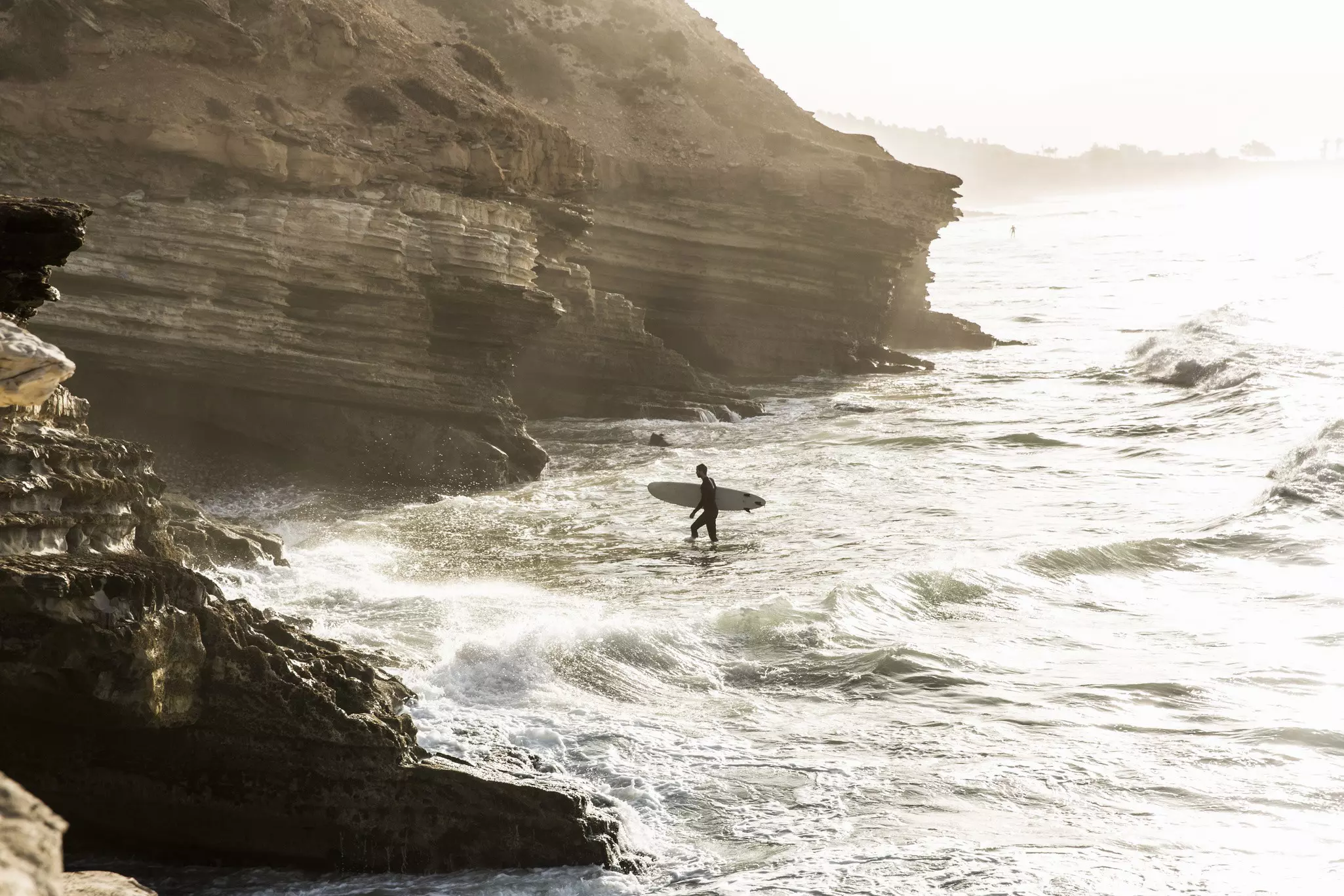 Surfer walking toward the rocky shore with his surfboard during sunrise, Taghazout Bay, Morocco
