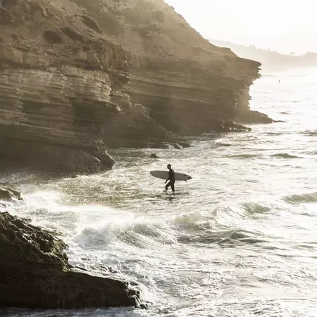 A surfer walks toward the rocky shore with his surfboard during sunrise.