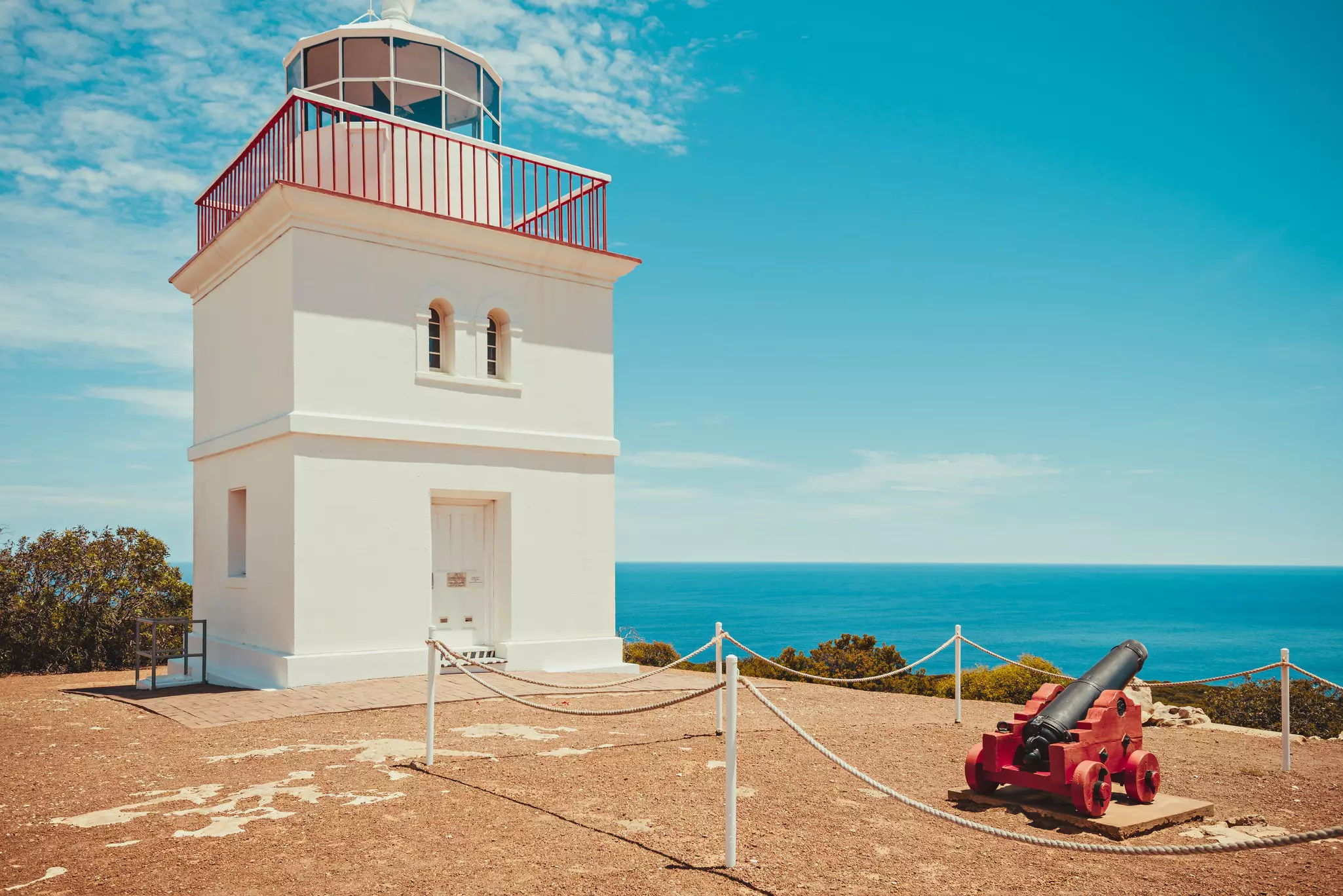 A low-level square lighthouse on a cliff top near an old canon pointing out at sea.