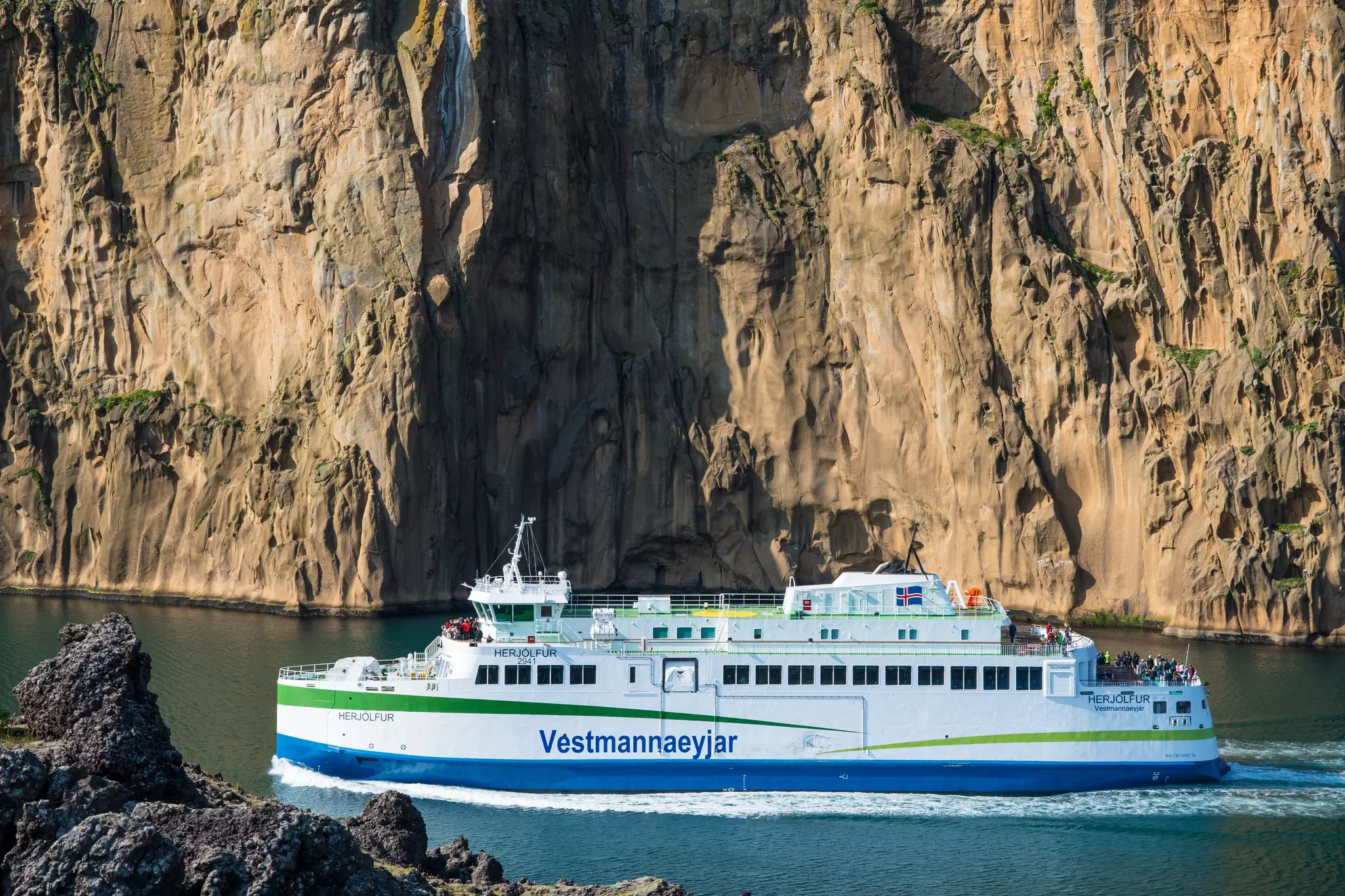 Large white ferry with blue and green stripes and "Vestmannaeyjar" written on the side in the water between small rock outcroppings and a large rocky mountain beyond on a sunny day.