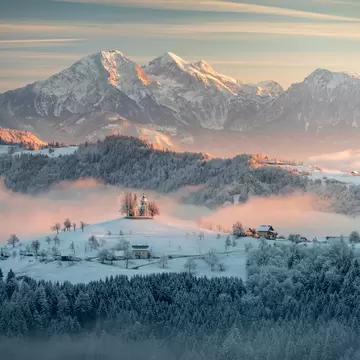 A foggy sunrise in a snowy countryside with a church on a hill.