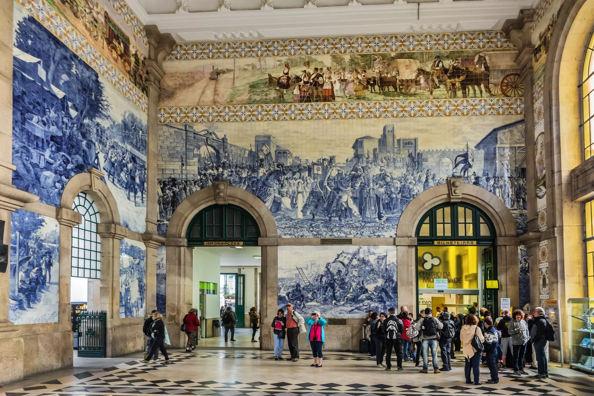 Tourist look at and photograph the blue and white tiled walls of a main hall in a train station that show historical scenes of Portuguese history.