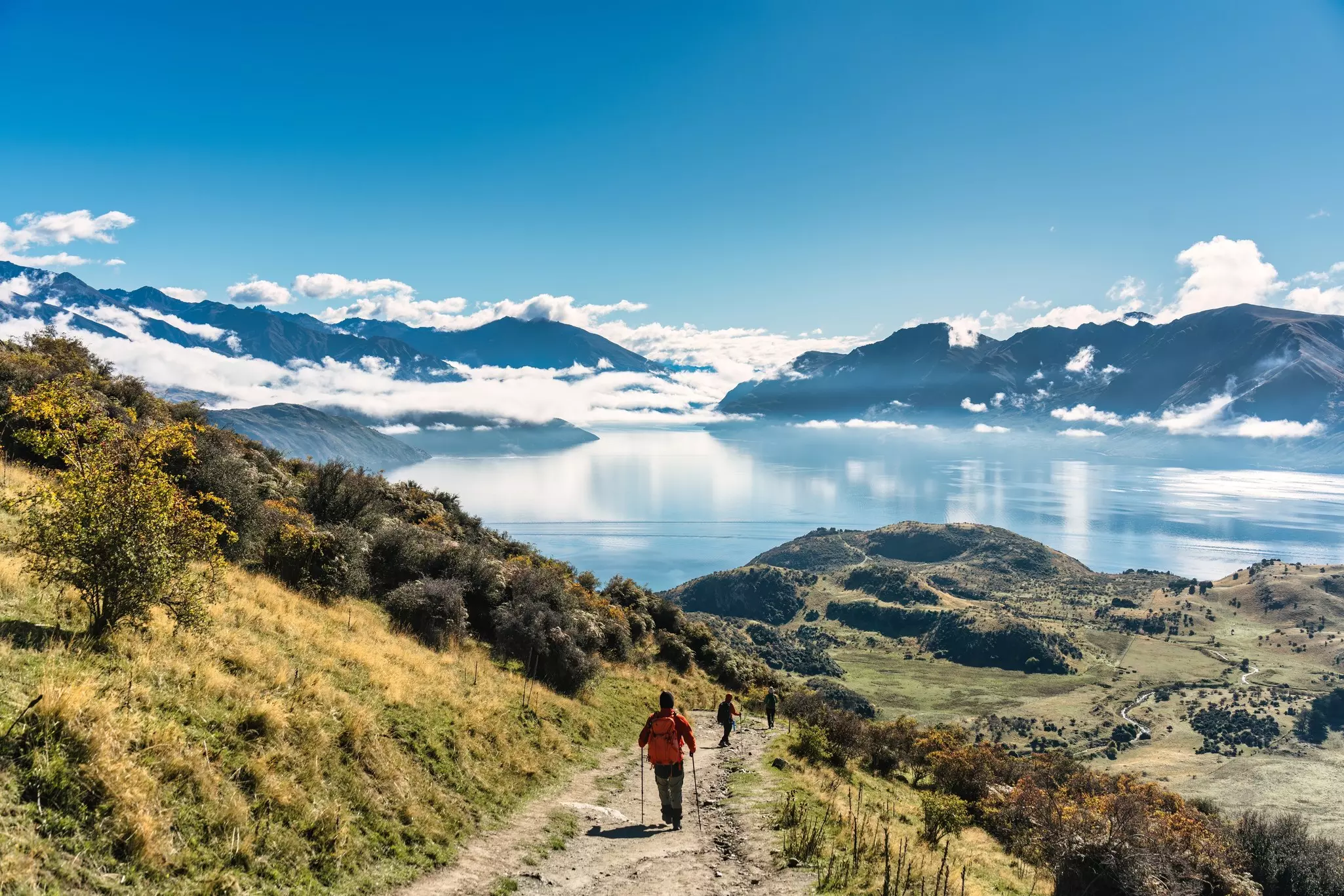 Three hikers descend a trail that leads down towards a lake surrounded by mountains.