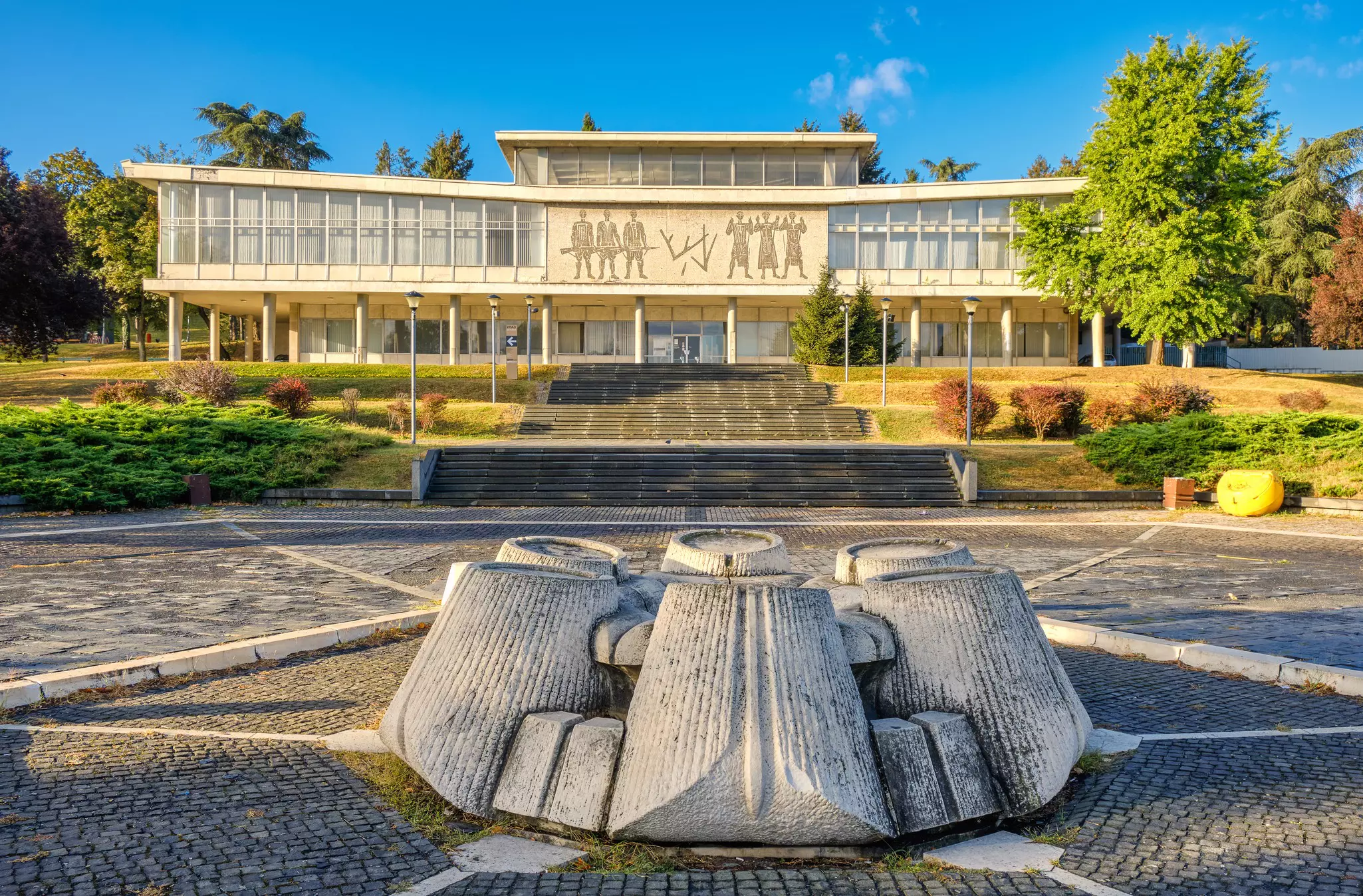 An abstract concrete sculpture centers a cobbled plaza in front of a white museum building with narrow columns and mosaic figures on its facade.