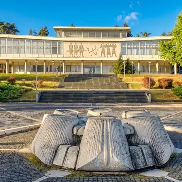 An abstract concrete sculpture centers a cobbled plaza in front of a white museum building with narrow columns and mosaic figures on its facade.