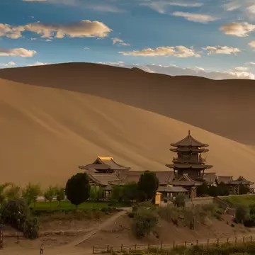 Multi-tiered Chinese pagoda and temple buildings among trees at the foot of a huge sand dune near Dunhuang, China.