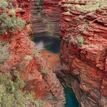 Deep canyon of stacked red rock ledges with a dark pool and small figures on a sandy bank below, Karijini, Western Australia