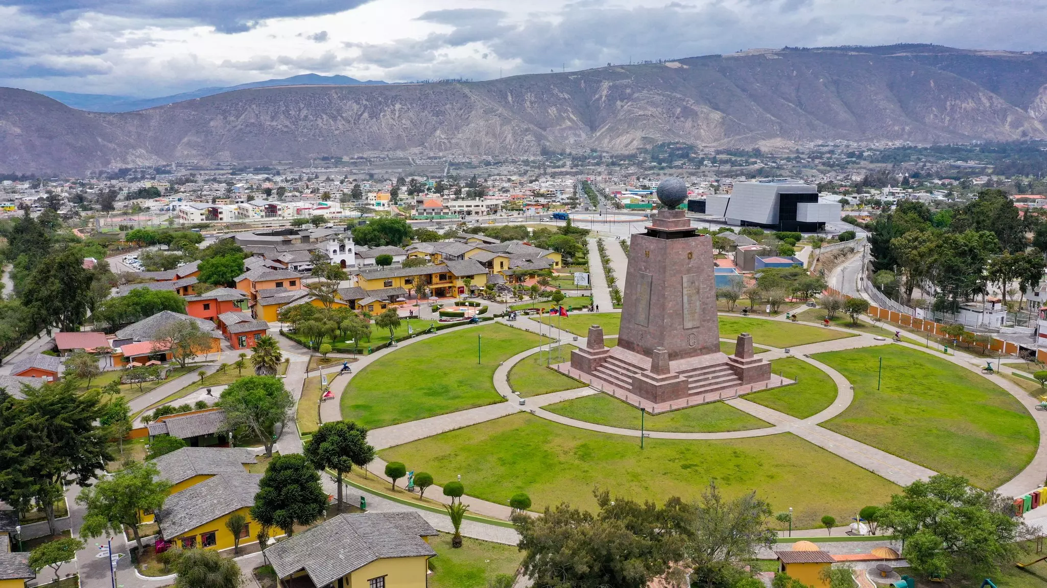 An aerial view of a large stone monument on a grass lawn in a city. Mountains are visible in the distance.