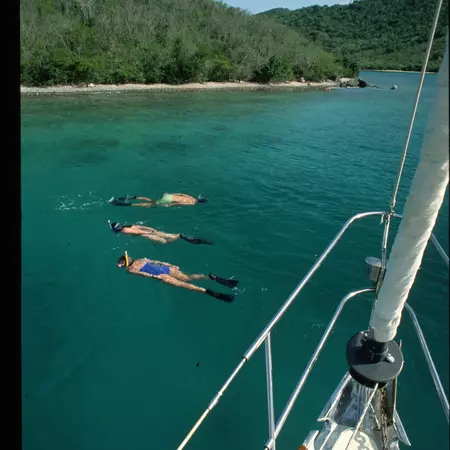 Three people snorkel in blue-green water beyond a sailboat's prow on a sunny day.