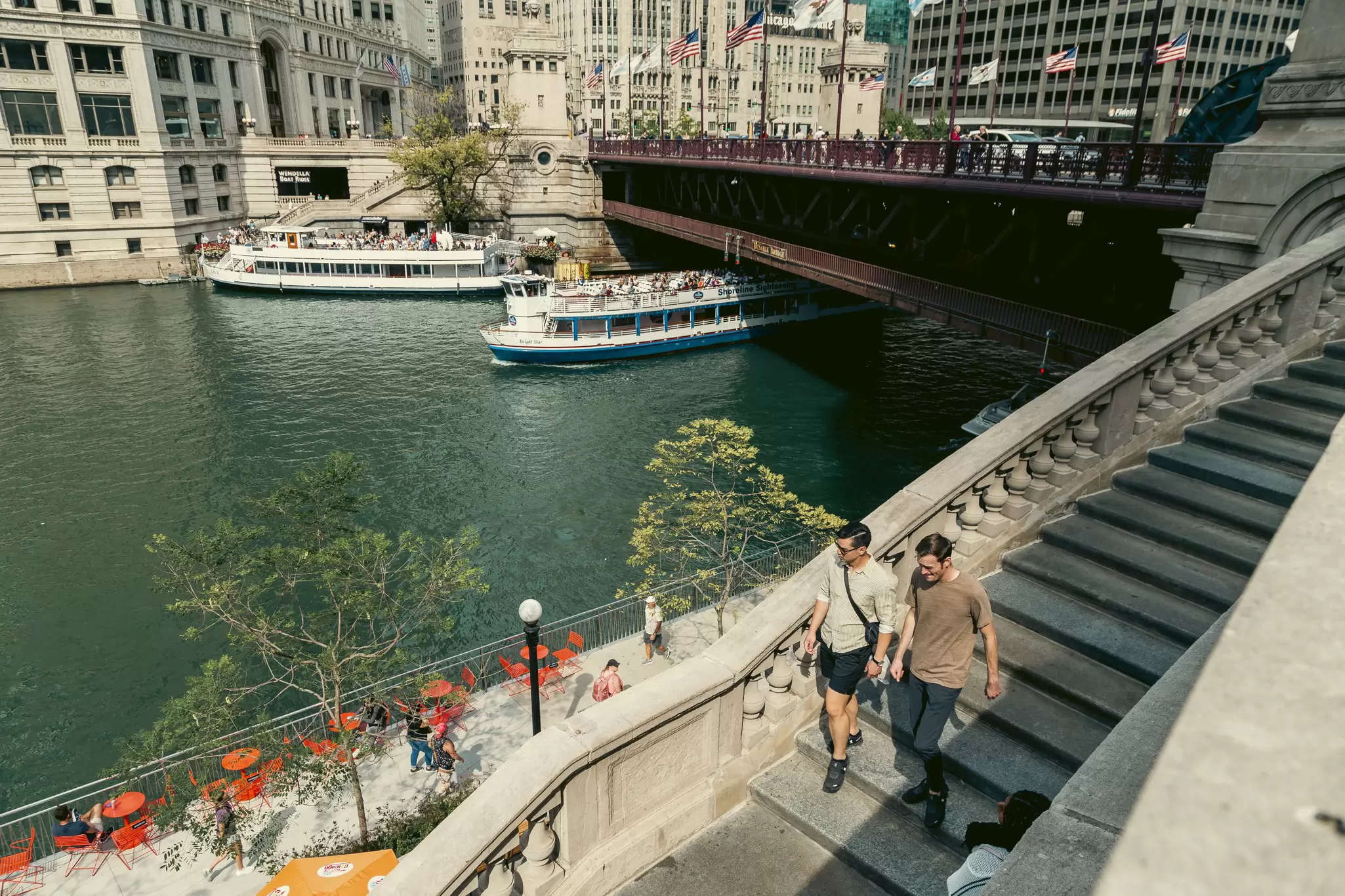 Tour boats coming out from under a bridge as people walk along the pathway next to the water