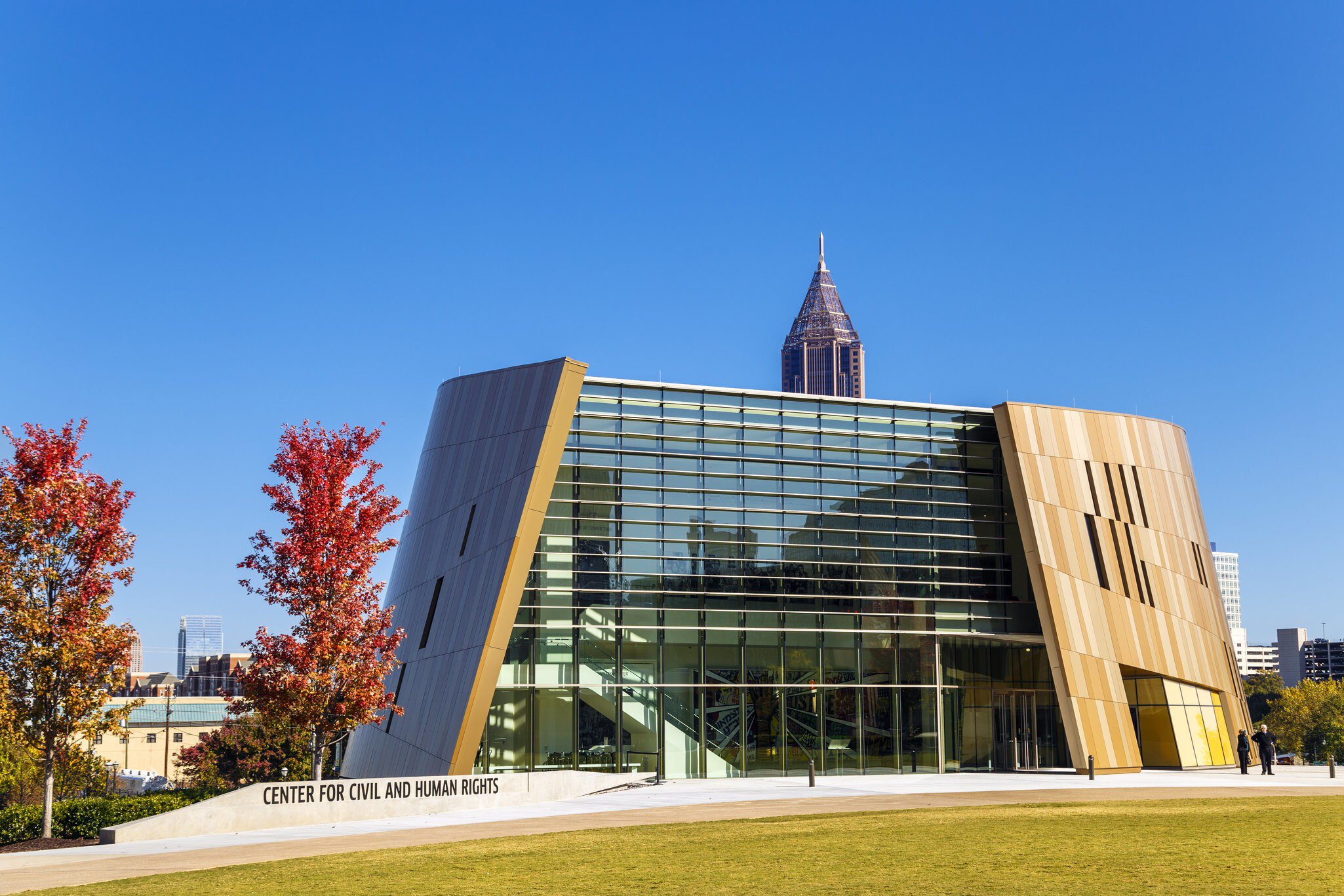 An angular glass and bronze-clad building behind red-leaved trees, a spired tower rising beyond.