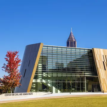 An angular glass and bronze-clad building behind red-leaved trees, a spired tower rising beyond.