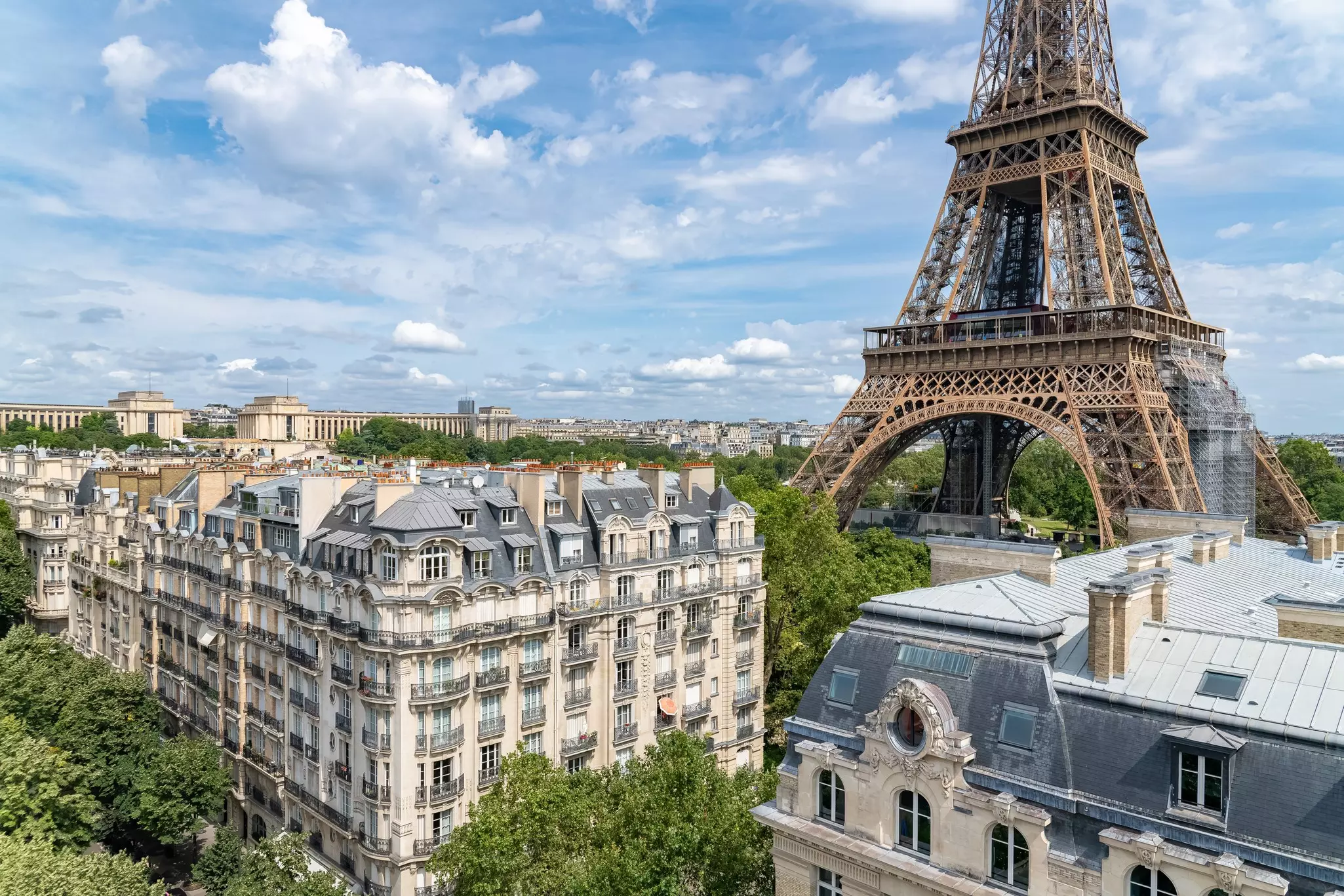 The base of the Eiffel Tower in Paris, with nearby buildings in the foreground; green trees surround the structures.