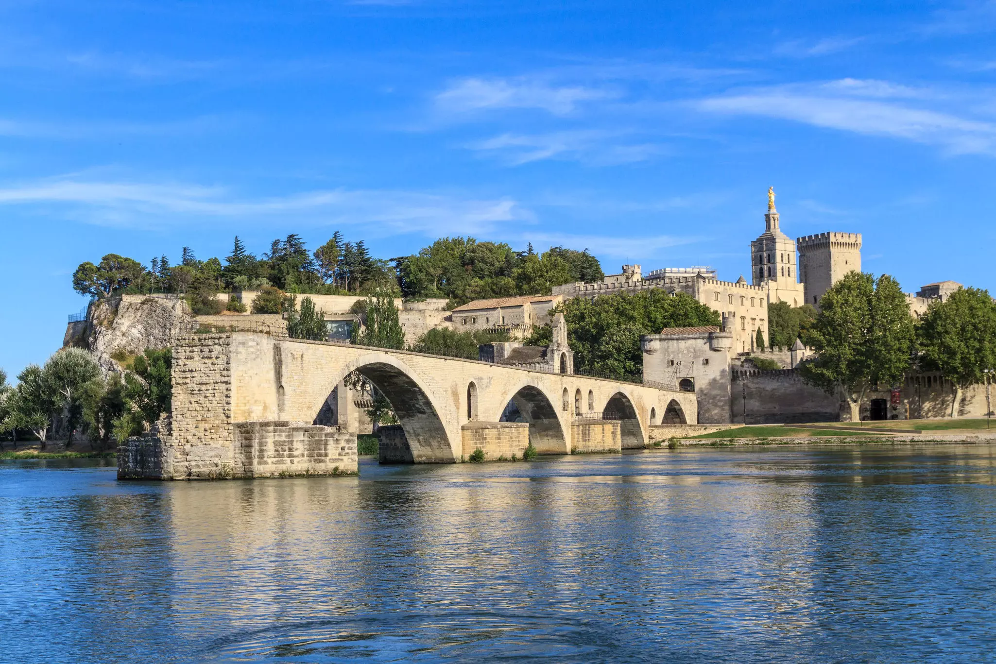 A ruined stoned bridge with arches juts into a river. Historic buildings stand on shore behind the bridge.