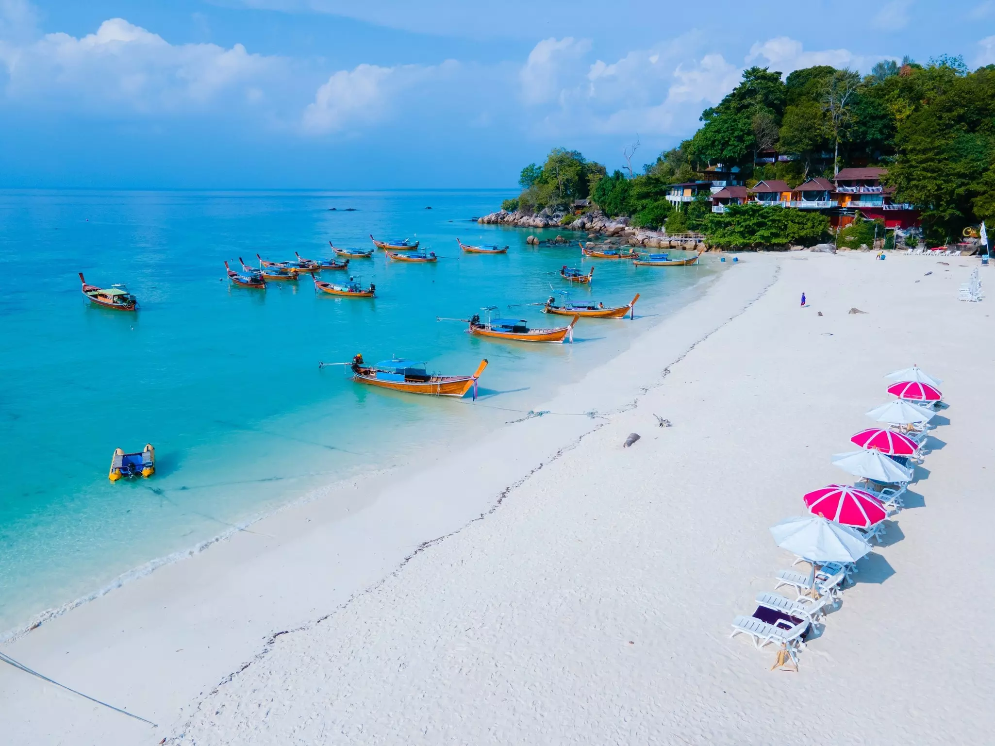 A line of chaise lounges with umbrellas sits on a wide white sand beach. Small fishing boats are moored in the shallow water just offshore.