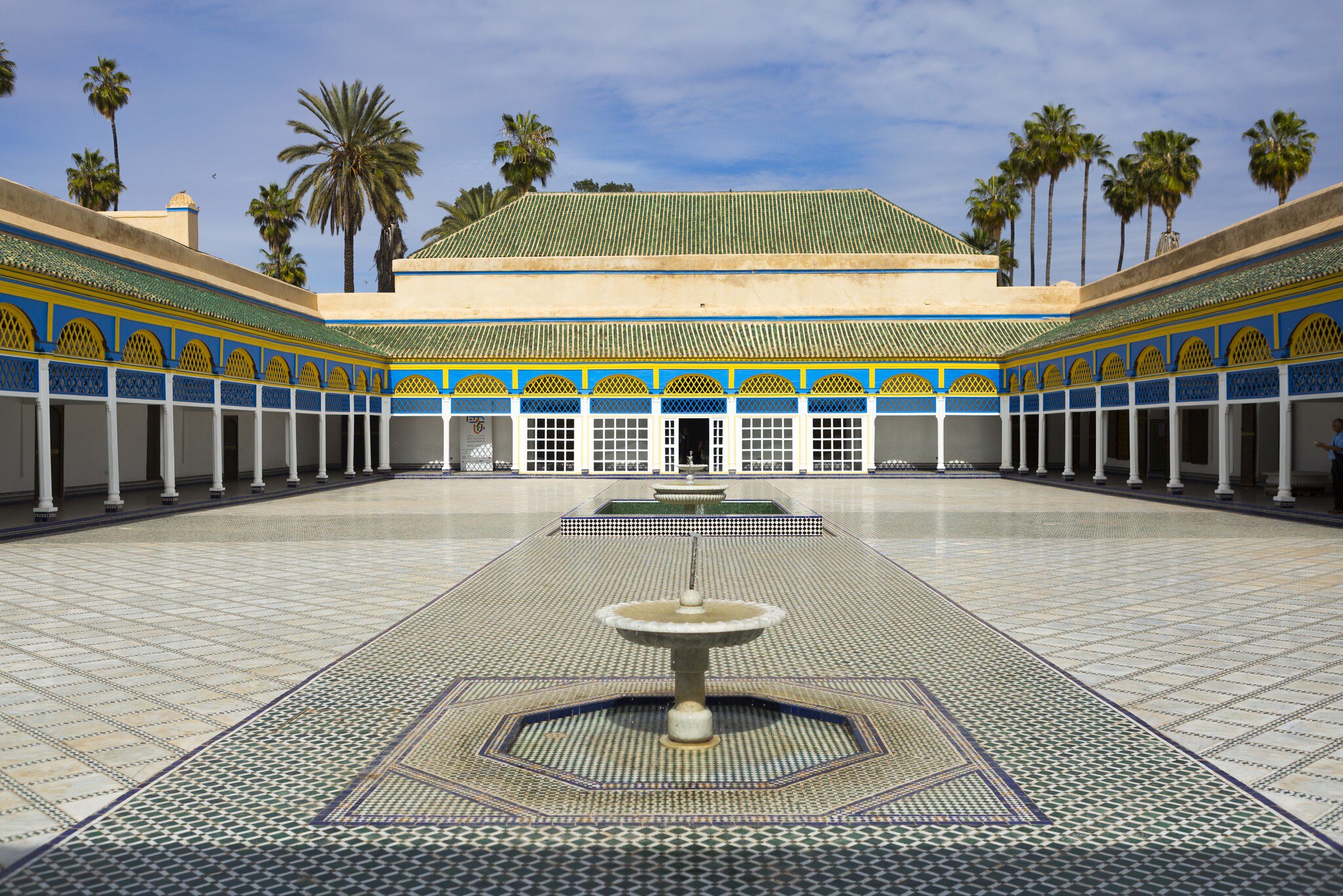 Empty tiled courtyard with a low marble fountain, blue-and-yellow painted arcades on white columns, palms above.