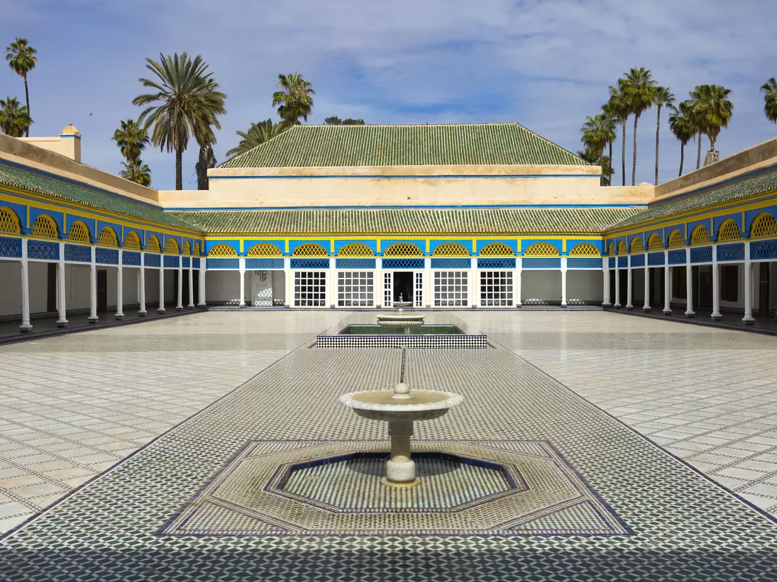 Empty tiled courtyard with a low marble fountain, blue-and-yellow painted arcades on white columns, palms above.