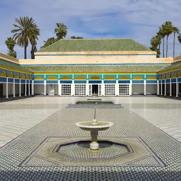 Empty tiled courtyard with a low marble fountain, blue-and-yellow painted arcades on white columns, palms above.