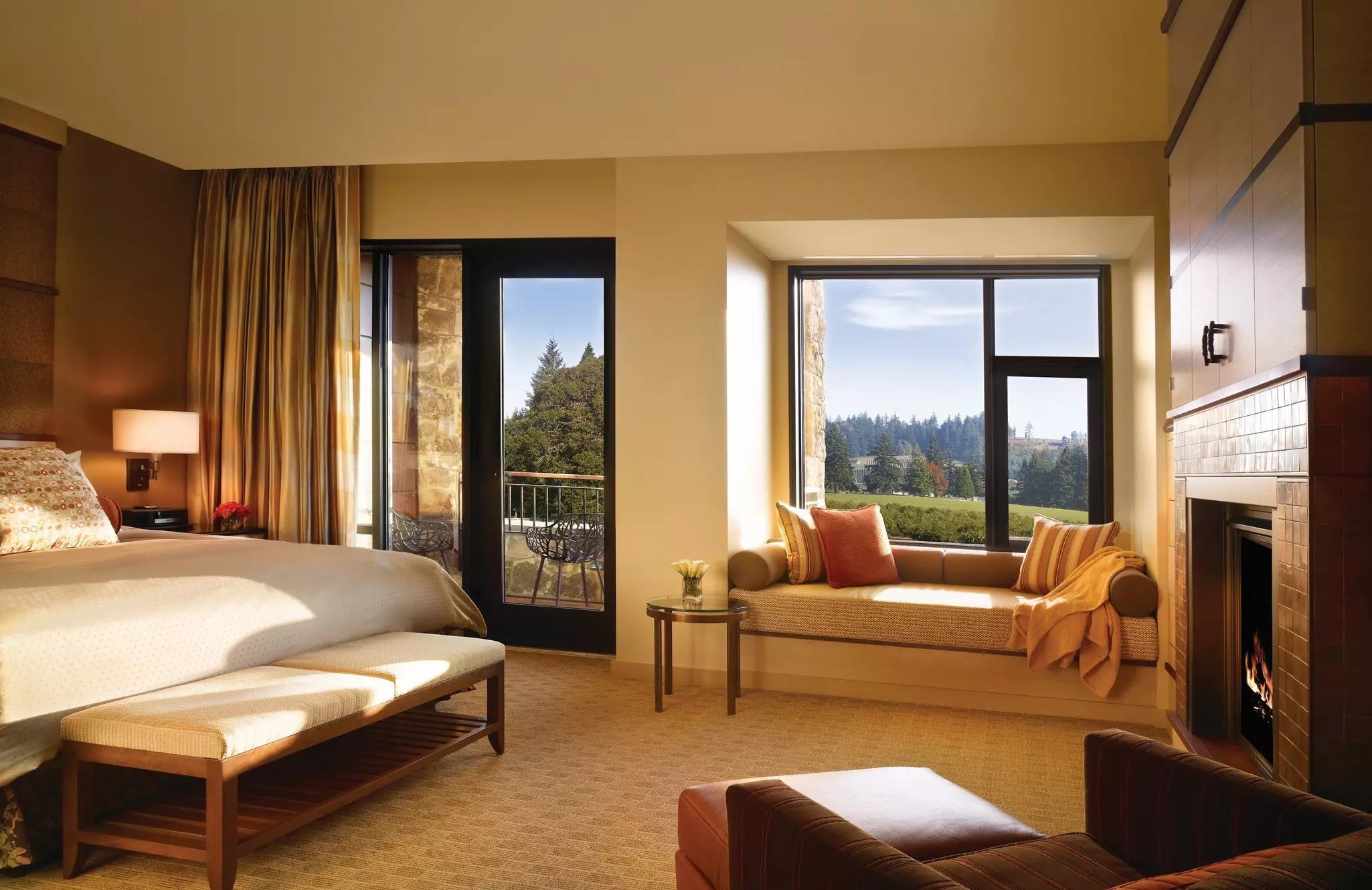 A king-sized bed with white coverlet in a hotel room with a wall of windows looking out over trees and green space.