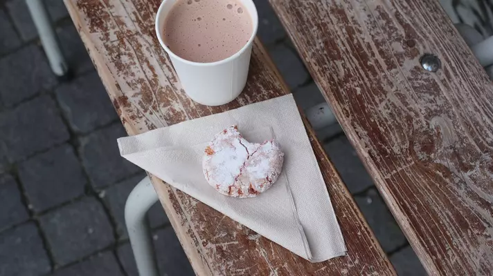 Hot chocolate in a paper cup and a cookie on a folded paper napkin rest on a bench.