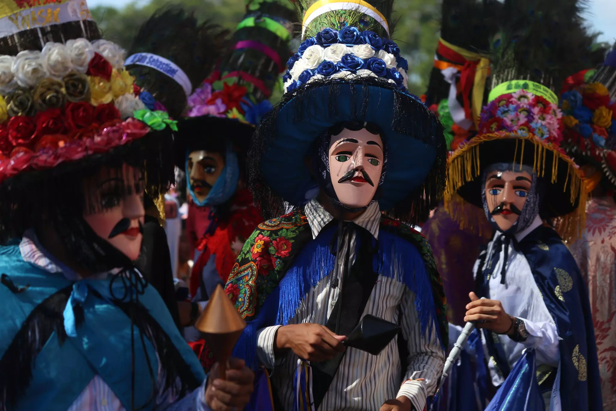 People in costumes with elaborate hats decorated with flowers. Each one wears a mask that fully covers their face.