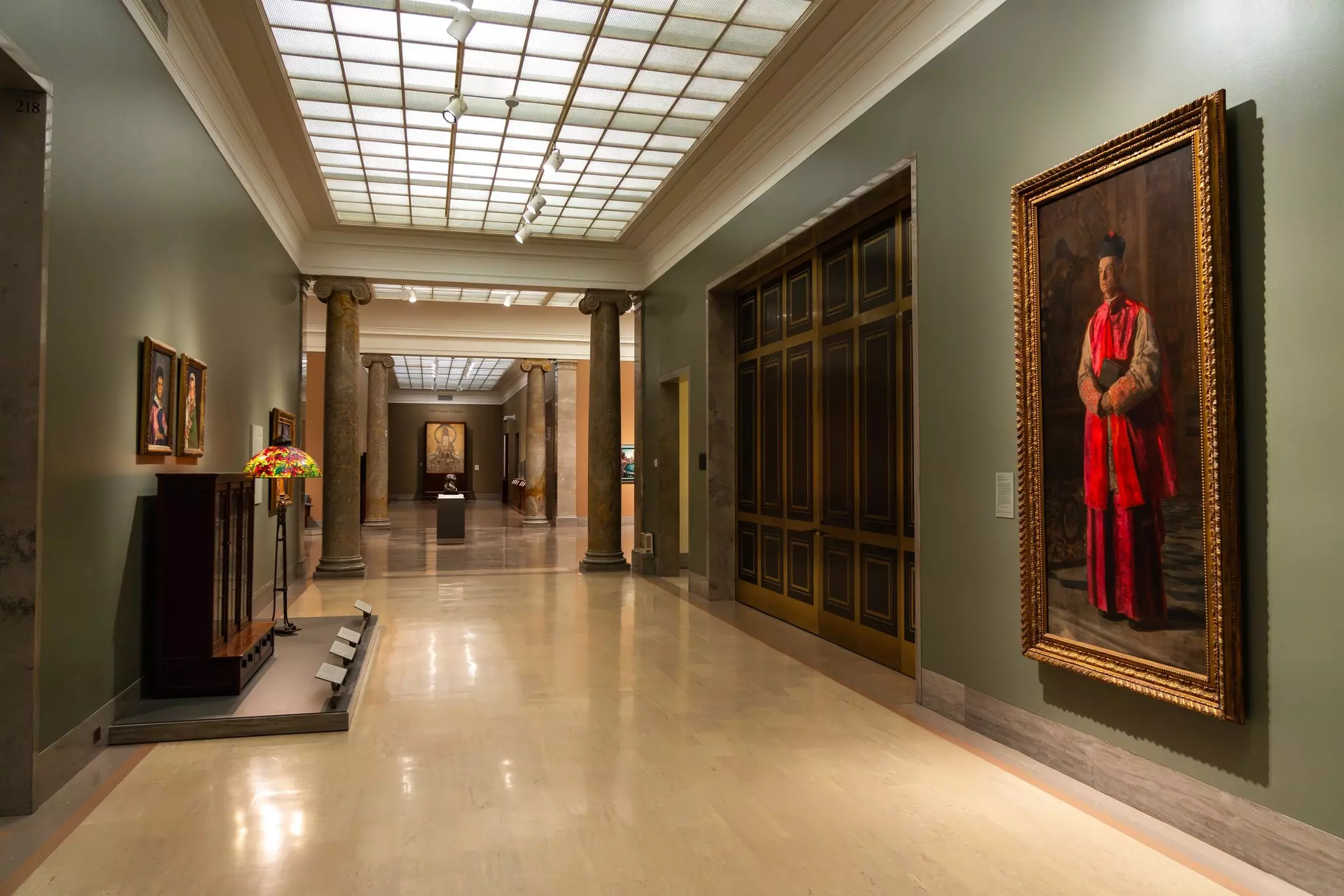 Paintings and decorative arts works are on display in a corridor of an art museum.
