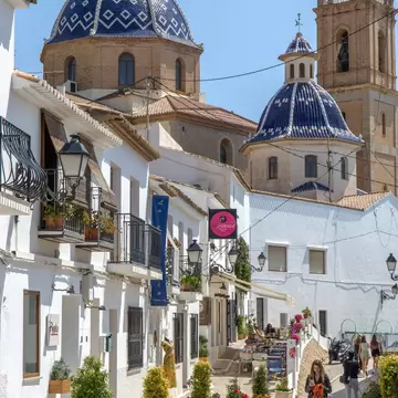 Sloping street of white houses with potted flowers leading to two blue and white tiled church domes in Altea.