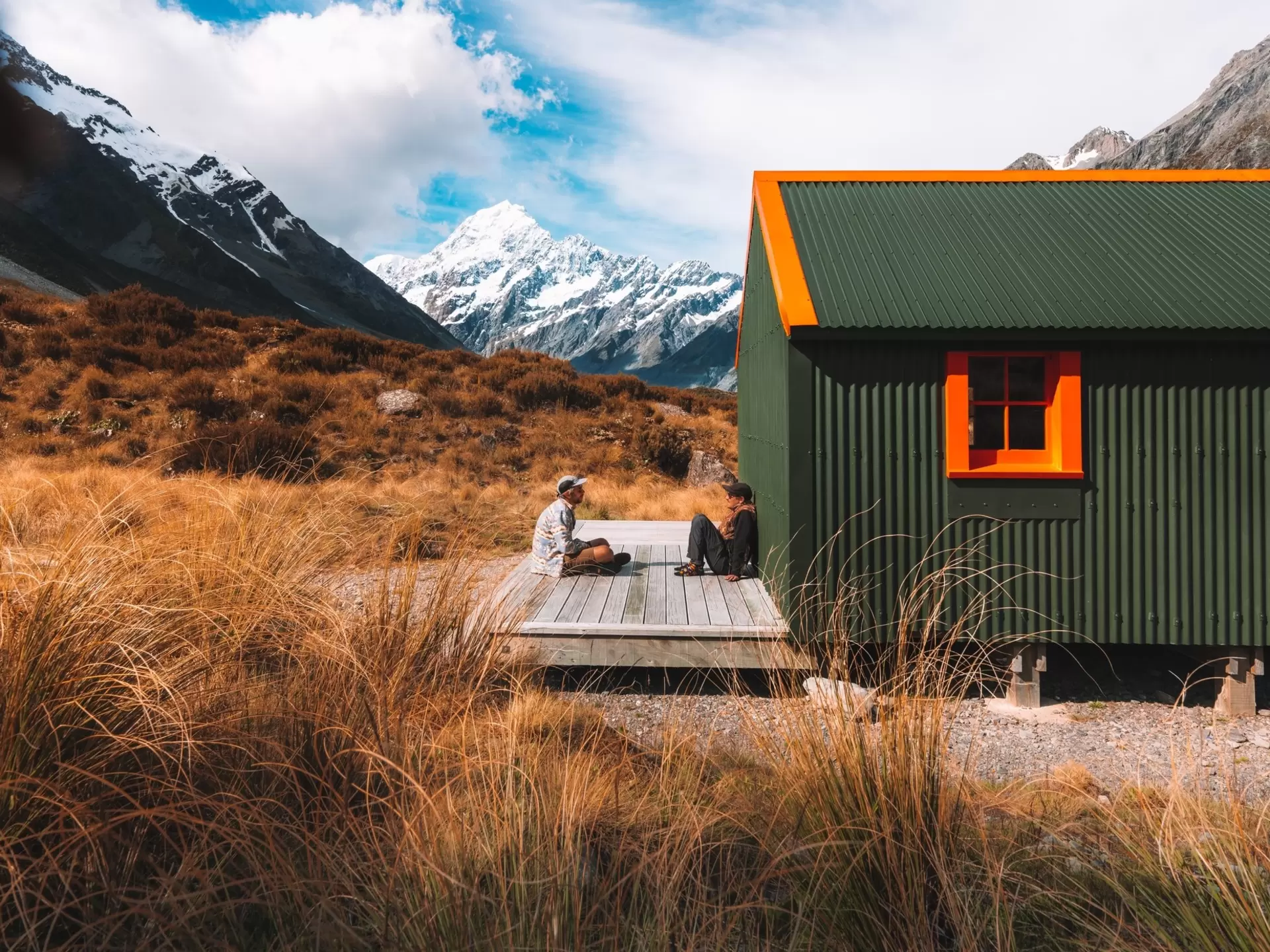 Two men sitting on the porch in front of a green hut with mountains in the background.