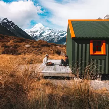 Two men sitting on the porch in front of a green hut with mountains in the background.