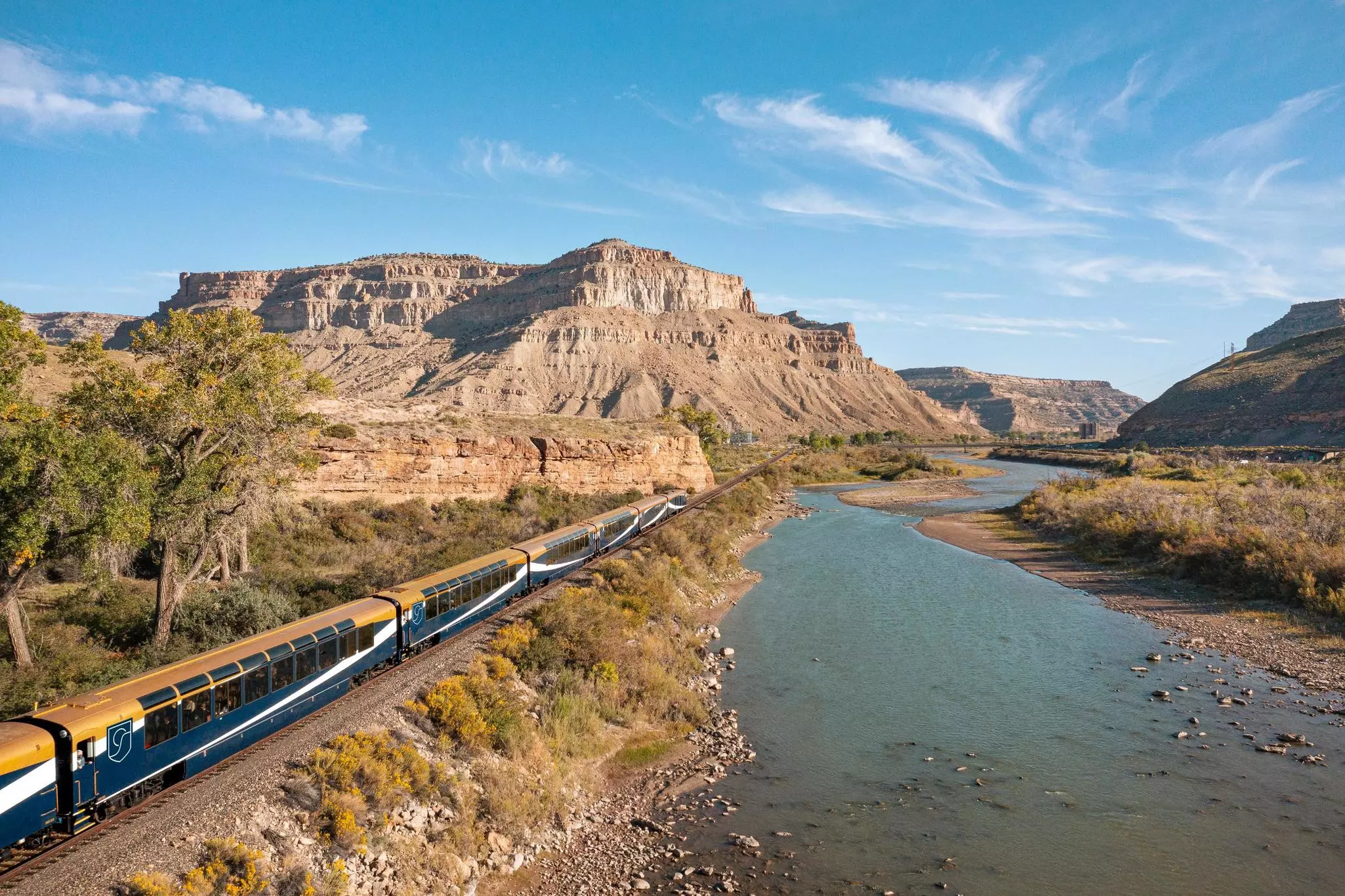 A blue and yellow train traveling beside a low river with stepped mountains in the distance on a sunny day.