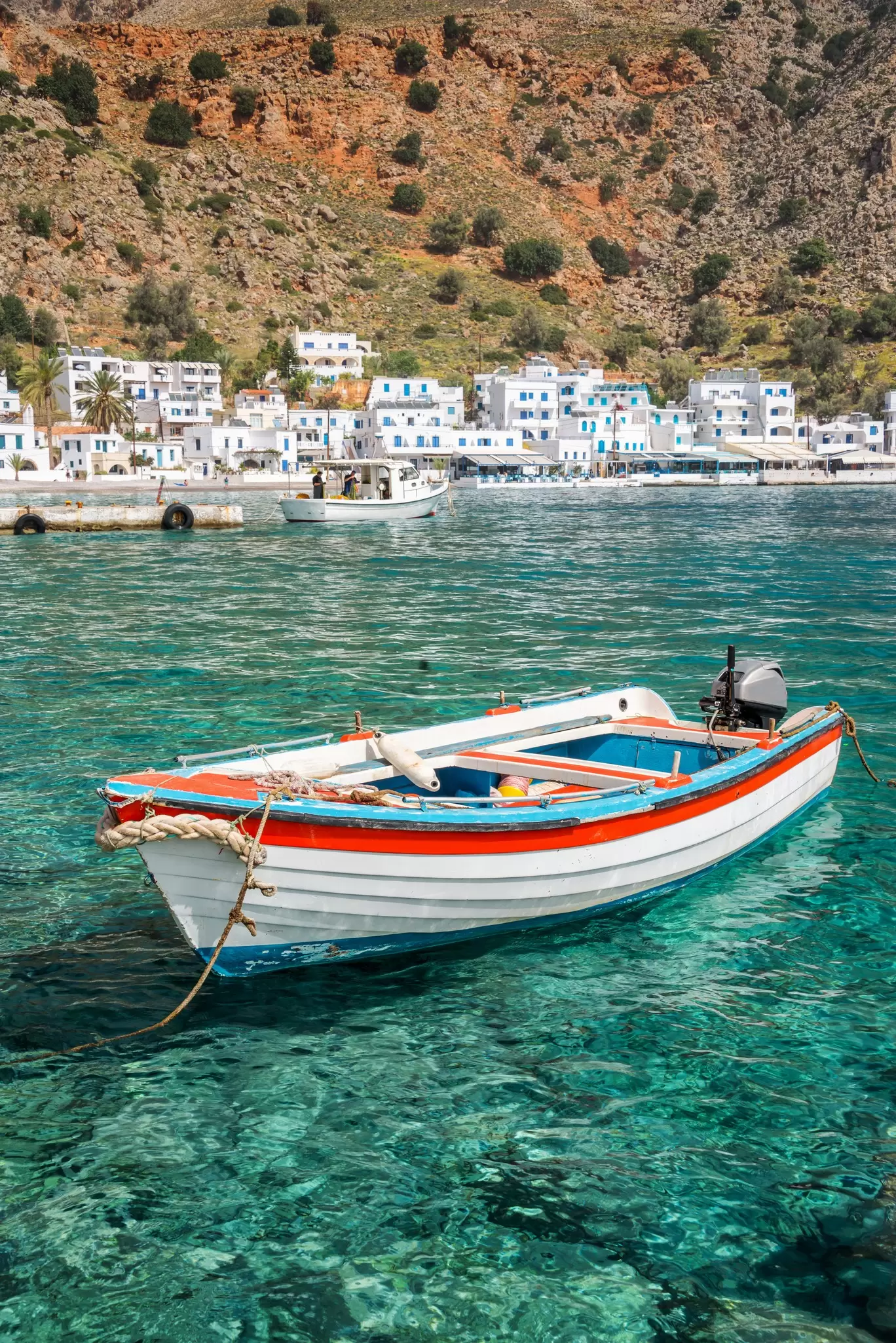 A fishing boat in turquoise water offshore a village with white buildings below a dry hillside.