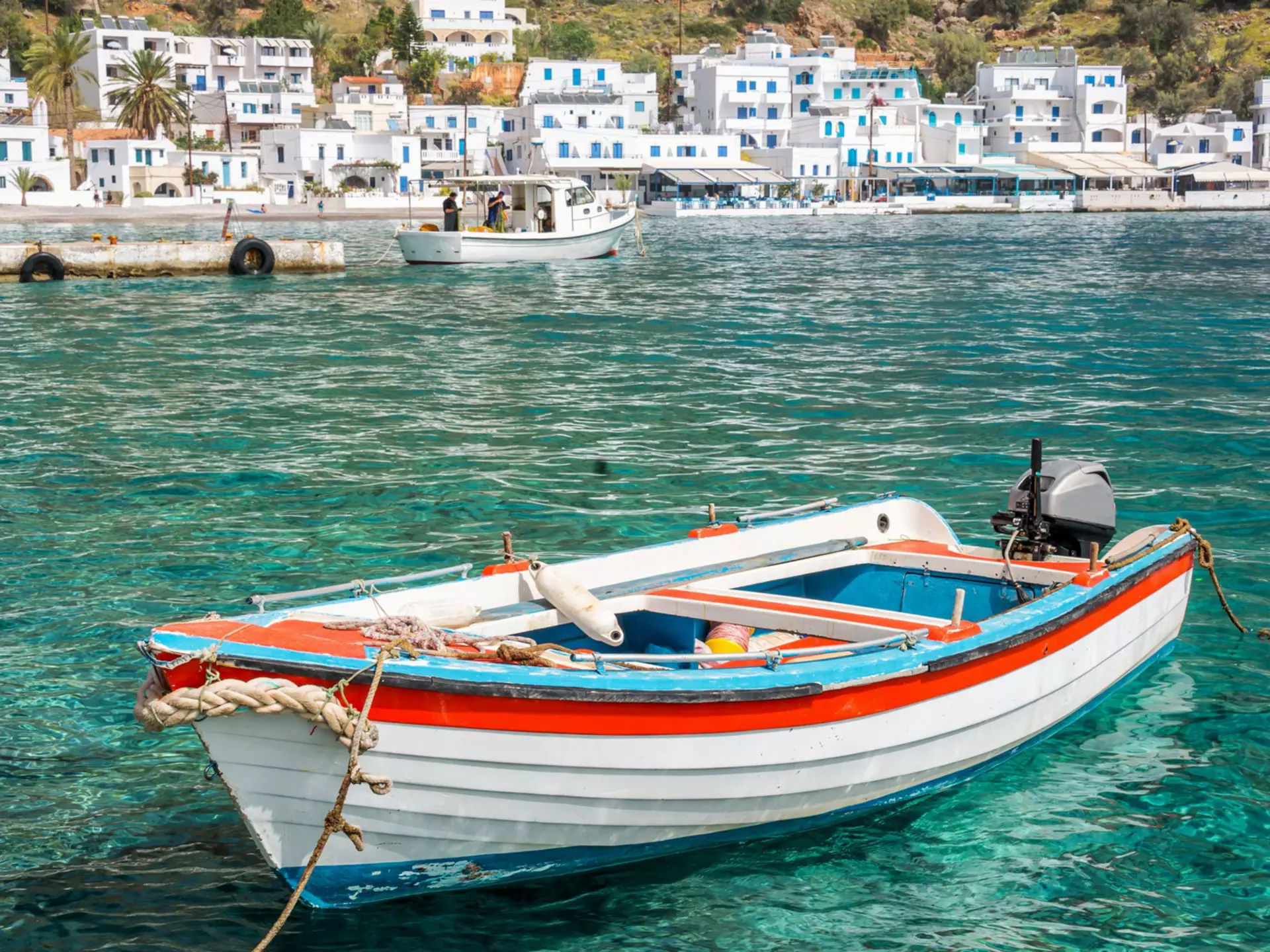 A fishing boat in turquoise water offshore a village with white buildings below a dry hillside.