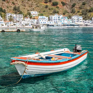 A fishing boat in turquoise water offshore a village with white buildings below a dry hillside.