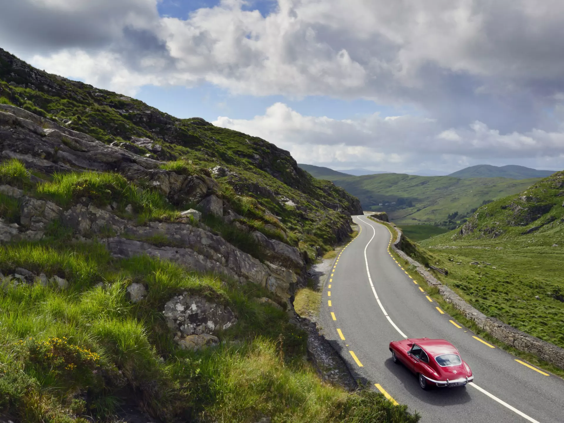An old-fashioned red car drives along a road set on rolling green hills under a cloudy sky