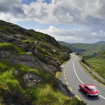 An old-fashioned red car drives along a road set on rolling green hills under a cloudy sky