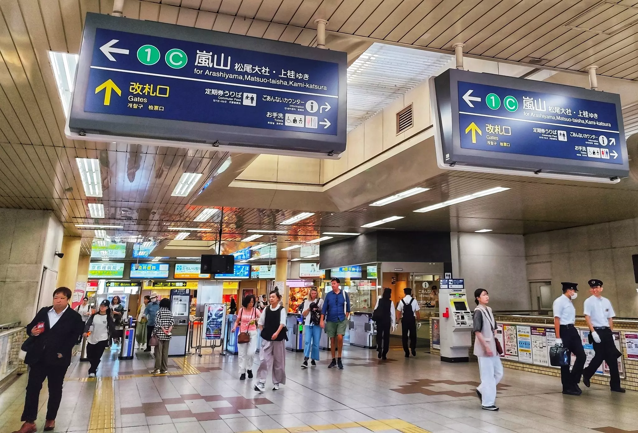 People walking in a subway station near the turnstiles. Big blue directional signs in Japanese and English hang overhead