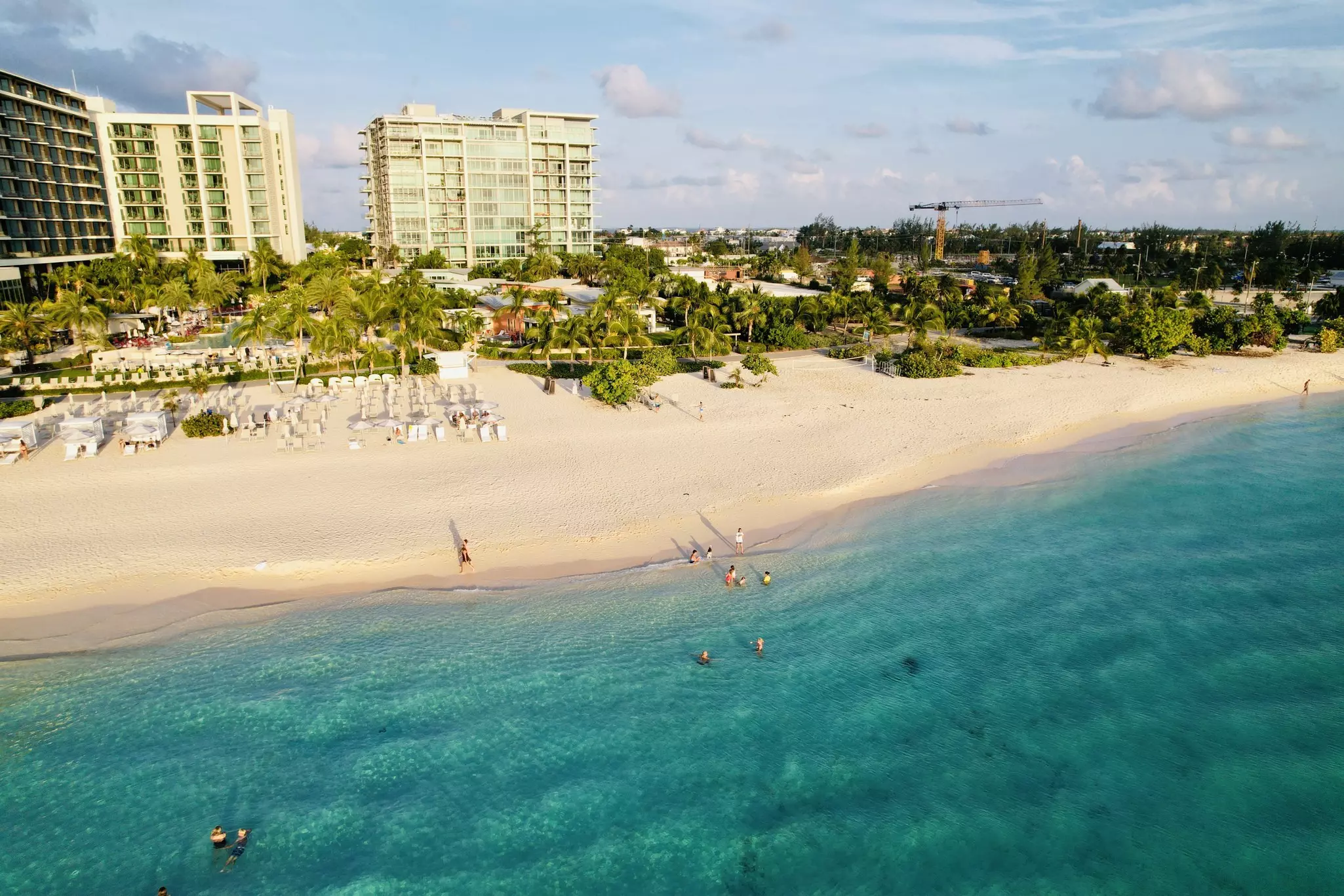 People swim in the blue ocean that laps at the edge of a sandy beach backed by a hotel resort.