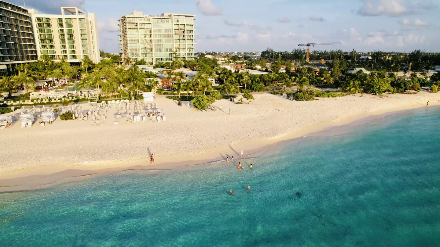 People swim in the blue ocean that laps at the edge of a sandy beach backed by a hotel resort.