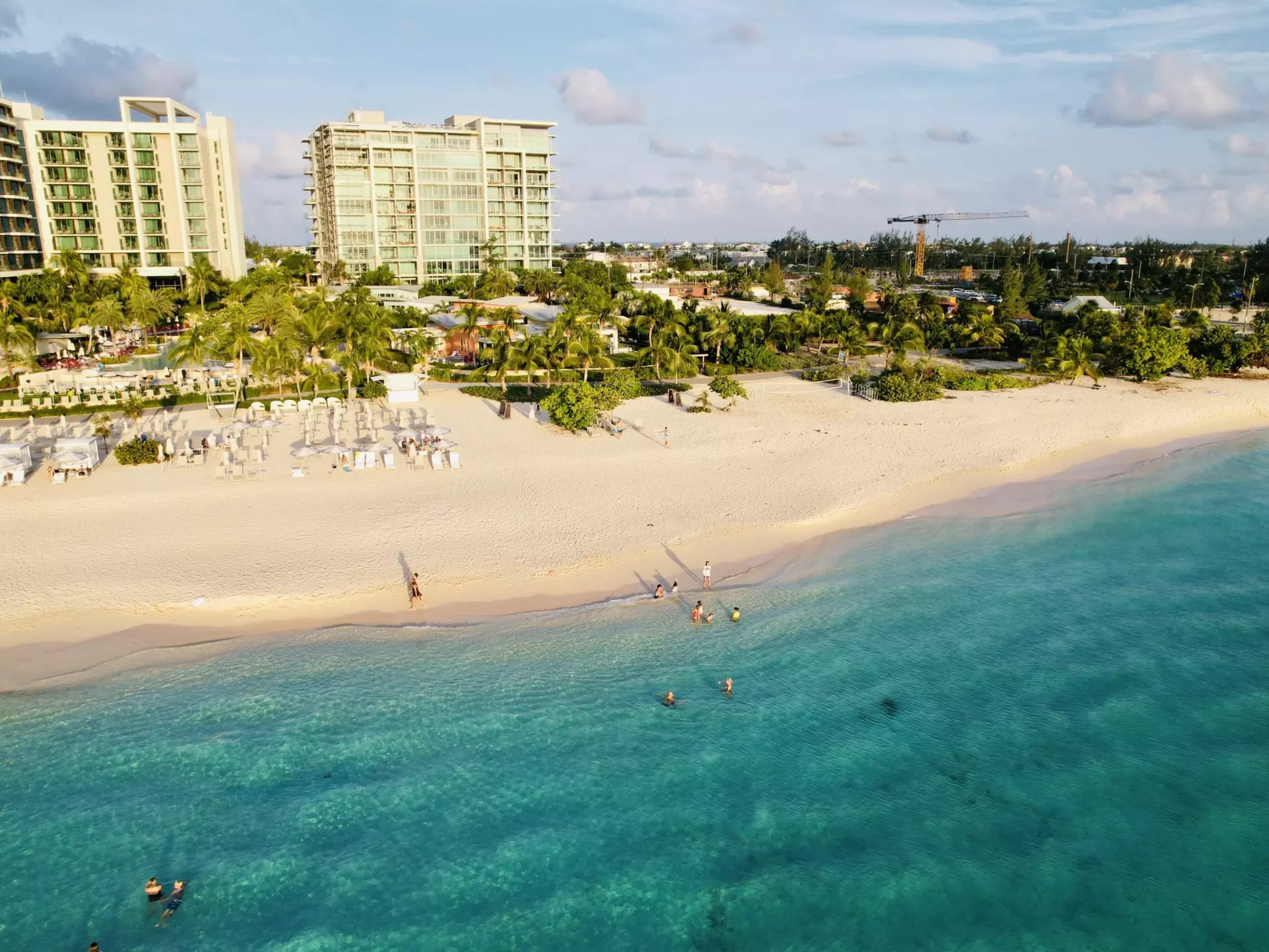 People swim in the blue ocean that laps at the edge of a sandy beach backed by a hotel resort.