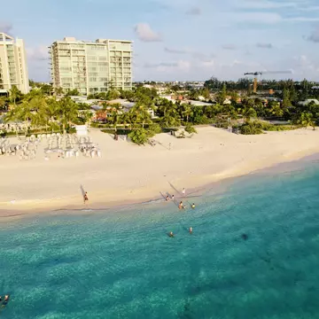 People swim in the blue ocean that laps at the edge of a sandy beach backed by a hotel resort.