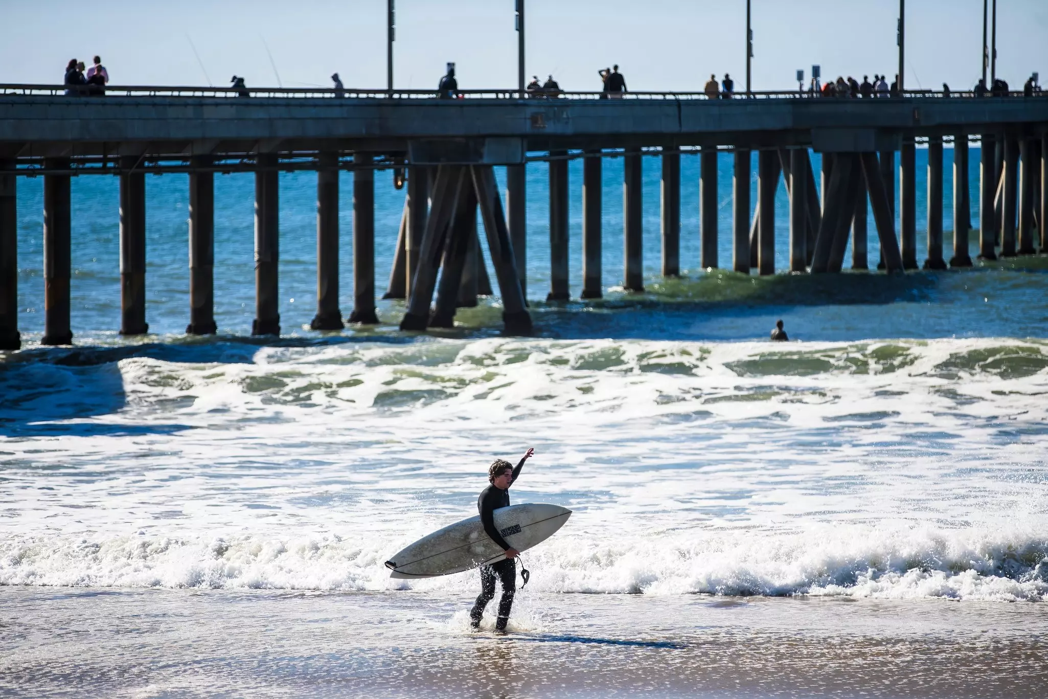 A surfer stands in the foam looking out at a rainbow on the horizon