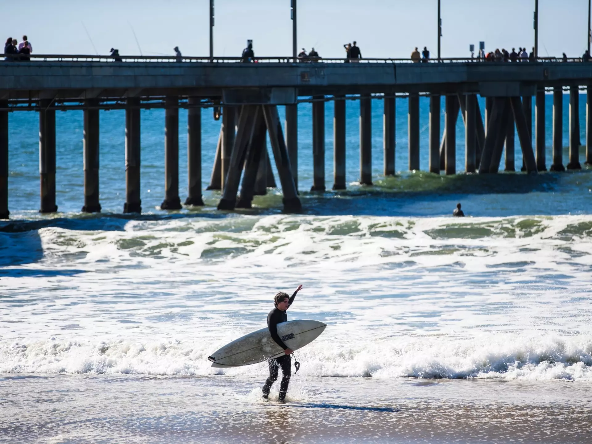 A surfer stands in the foam looking out at a rainbow on the horizon
