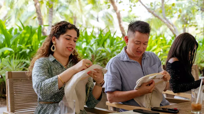 A man and woman sit in front of lush greenery with embroidery hoops in their hands.