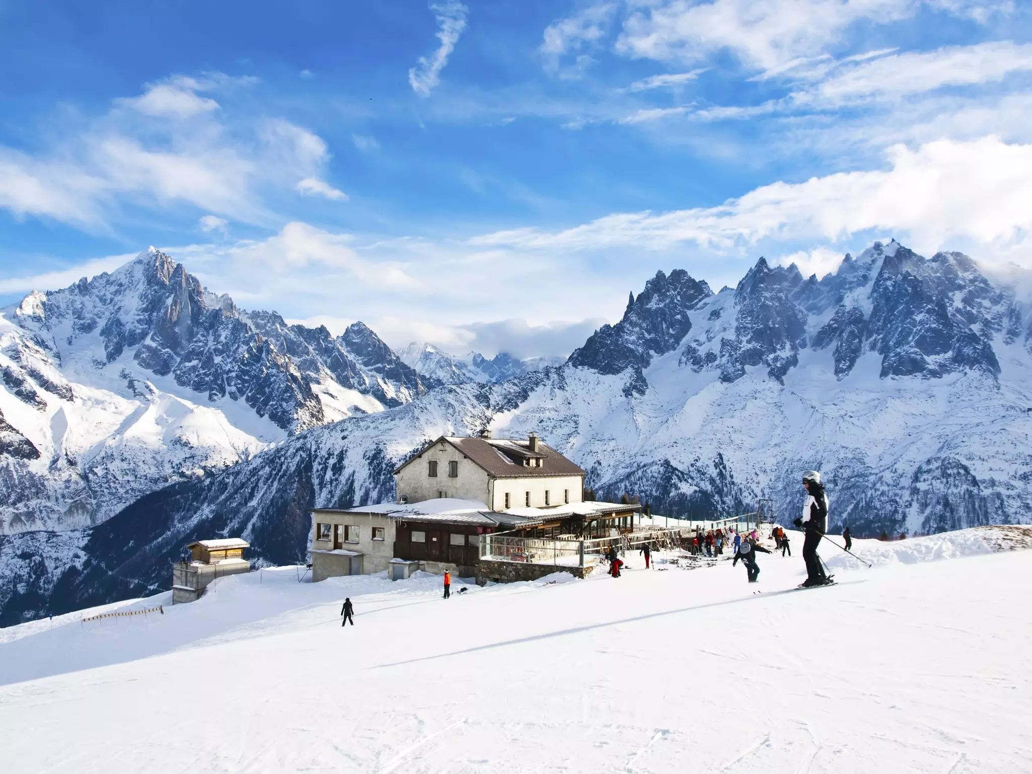 People ski past a lodge on a slope at a resort, with jagged peaks visible in the distance.