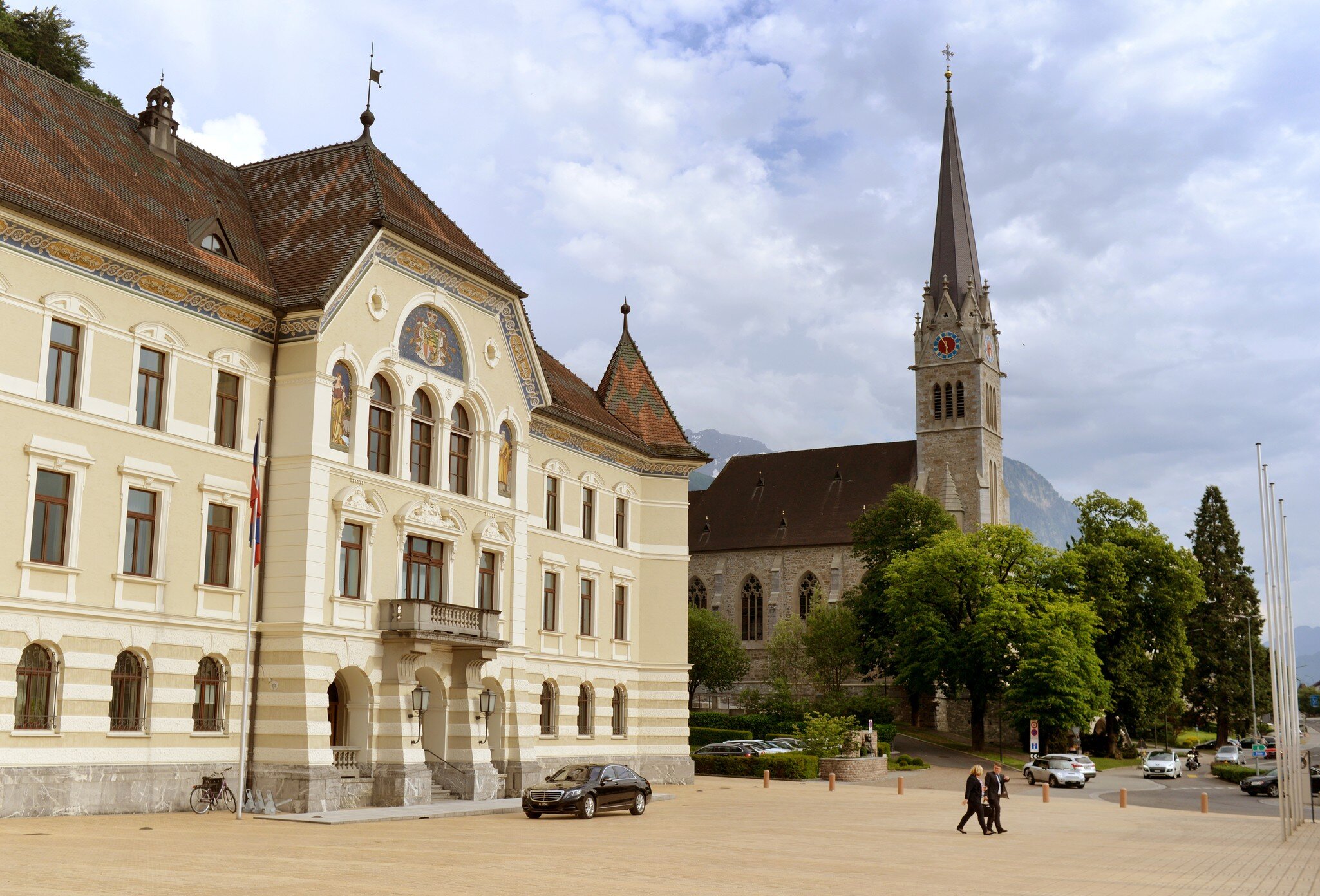 Vaduz's Government House beside the Gothic Revival spire of St Florin Cathedral, with mountains behind