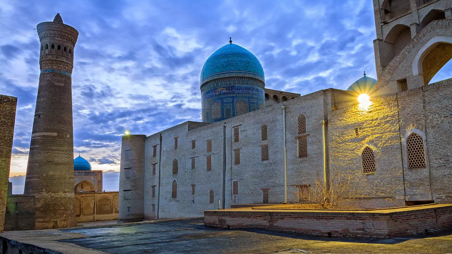 The blue dome and brown mud structures of the Kalon Mosque and minaret under cloudy, dusk skies.