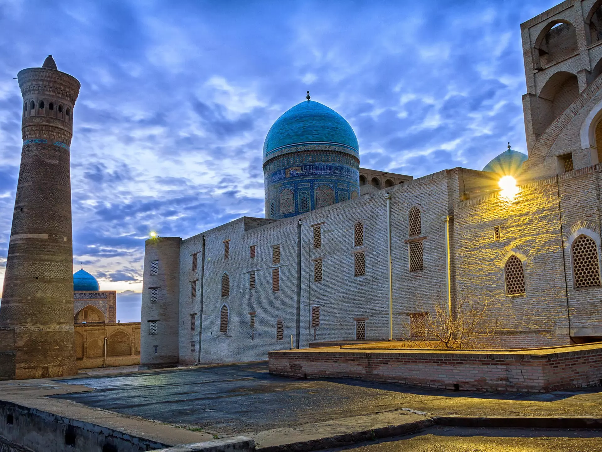 The blue dome and brown mud structures of the Kalon Mosque and minaret under cloudy, dusk skies.